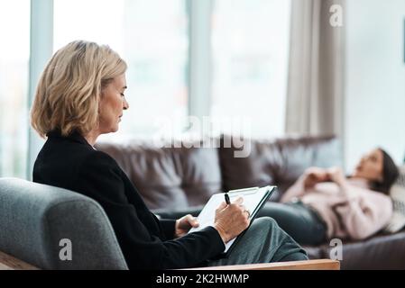 Parlez-moi de votre enfancePrise de vue d'un psychologue mature qui écrit des notes au cours d'une séance thérapeutique avec sa patiente. Banque D'Images