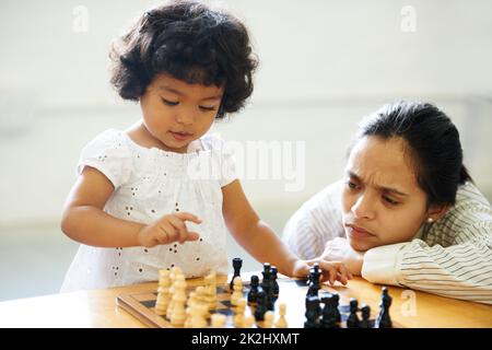 Quand a-t-elle eu cette intelligence ?Une petite fille mignonne jouant aux échecs pendant que sa mère regarde dessus. Banque D'Images