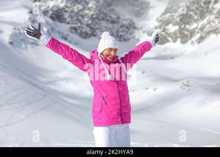 Jeune femme en veste de ski rose, chapeau et gants d'hiver, les bras au-dessus de propagation, souriant, heureux à la montagne couverte de neige derrière elle. Banque D'Images