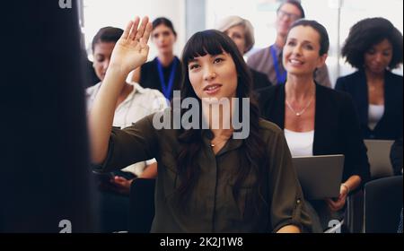 J'ai tant de questions. Photo courte d'une jeune femme d'affaires attirante assise avec ses divers collègues et levant la main au bureau. Banque D'Images