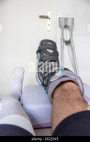 L'homme européen après l'opération de rupture de tendon d'Achille est de retour à la maison avec des chaussures spéciales de physiothérapie et des béquilles pour la récupération à la maison avec la médecine saine analgésique médicaments pilules contre la jambe blessant Banque D'Images