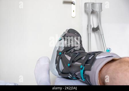 L'homme européen après l'opération de rupture de tendon d'Achille est de retour à la maison avec des chaussures spéciales de physiothérapie et des béquilles pour la récupération à la maison avec la médecine saine analgésique médicaments pilules contre la jambe blessant Banque D'Images