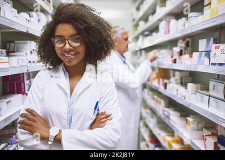 Nous avons ce dont vous avez besoin ici. Portrait d'une jeune pharmacienne gaie debout avec les bras repliés tout en regardant la caméra dans une pharmacie. Banque D'Images