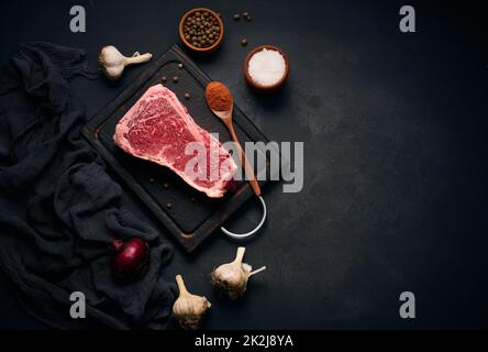 Morceau de viande de bœuf cru juteux sur l'os repose sur une planche à découper en bois, épices pour cuisiner sur fond noir. Filet de viande New York Banque D'Images