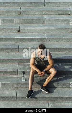 Homme sportif confiant assis sur des escaliers en béton après un entraînement physique à l'extérieur Banque D'Images