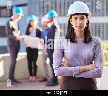 Des rénovations sont en cours. Photo d'une jeune femme d'affaires debout avec ses bras pliés et portant un casque de sécurité pendant que ses collègues se tiennent derrière elle. Banque D'Images