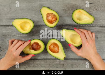 Vue sur table, femme mains avec ongles rouge holding avocats coupé en deux moitiés, de plus, certains d'entre eux à des semences autour sur bois gris 24. Banque D'Images