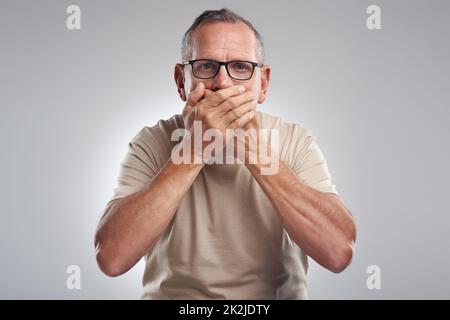 Je n'aurais pas dû le dire. Photo d'un homme mûr debout sur un fond gris dans le studio et couvrant sa bouche avec ses mains. Banque D'Images