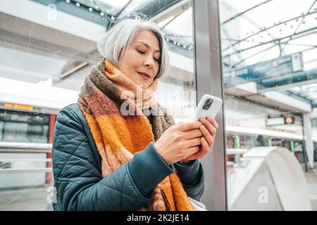 Une femme âgée envoie un message avec un téléphone portable et attend un train Banque D'Images