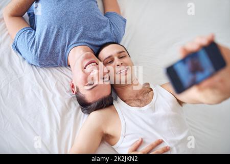 Faites-en un gardien avec un grand sourire. Un jeune couple gay prend un selfie tout en se relaxant au lit. Banque D'Images