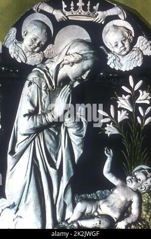École d'Andrea Della ROBBIA (1435-1525) l'adoration. La Vierge Marie, mains en position de prière, s'agenouille au-dessus du Christ naissant. En haut, les mains tiennent la couronne de la Vierge et deux chérubins regardent vers le bas. Au centre à droite se trouvent Madonna Lilies (Cilium candidum). Terre cuite émaillée. Banque D'Images