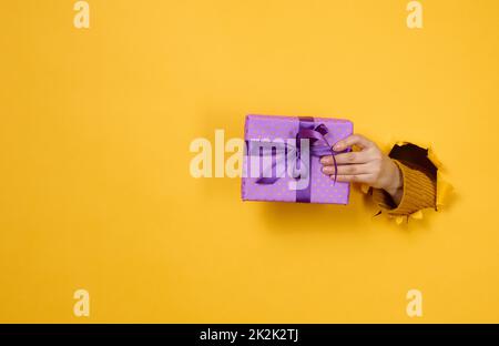 la main femelle tient une boîte avec un cadeau sur fond jaune, une partie du corps dépasse d'un trou déchiré dans un fond en papier. Félicitations, surprise de vacances. Banque D'Images