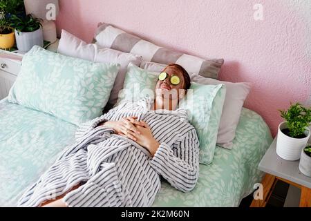 Masque et refroidissement. Prise de vue en grand angle d'une jeune femme attirante couchée sur son lit avec un masque de boue et des tranches de concombre sur son visage à la maison. Banque D'Images