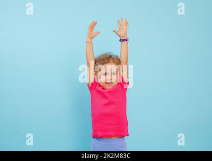 Portrait d'une jeune fille joyeuse aux yeux bleus avec des cheveux bouclés et équitables portant un T-shirt rose, levant les mains sur fond bleu. Banque D'Images