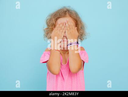 Portrait de la petite fille effrayée bouleversée couvrant les yeux avec les mains se cachant le visage sur fond bleu. Émotions, craintes, studio. Banque D'Images