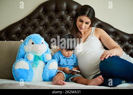 La technologie sans fil lui permet de se divertir partout. Prise de vue d'une femme enceinte regardant son petit garçon pendant qu'il joue avec un smartphone sur son lit. Banque D'Images