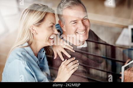 Un mariage heureux, une maison heureuse. Photo d'une femme mûre qui embrasse son mari alors qu'il utilise une tablette numérique sur le canapé à la maison. Banque D'Images