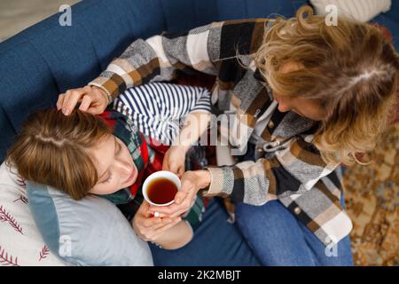 Femme donne du thé chaud à sa fille malade couché sur le canapé à la maison. Mère prend soin de sa fille malade Banque D'Images