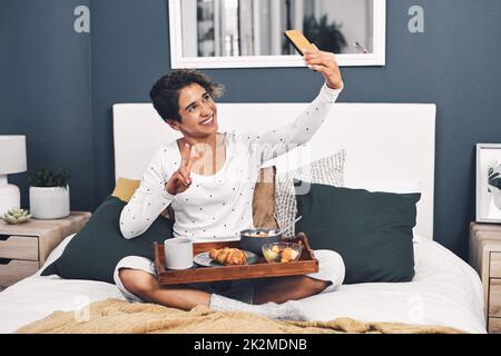 La vie d'un blogueur.Prise de vue en longueur d'une jeune femme attirante prenant un selfie en posant avec son plateau de petit déjeuner au lit. Banque D'Images