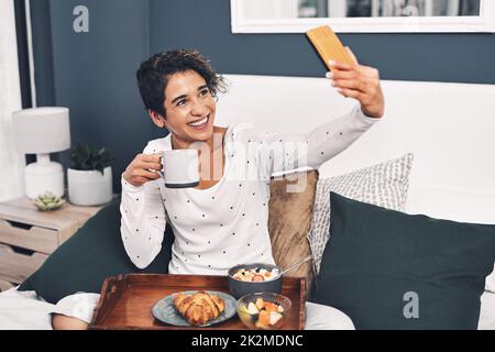 Heure du selfie.Prise de vue en longueur d'une jeune femme attirante prenant un selfie en posant avec son plateau de petit déjeuner au lit. Banque D'Images