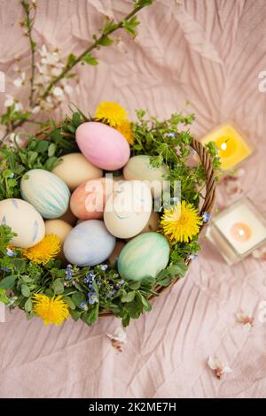 Sur un couvre-lit pastel est un panier d'herbe et de fleurs lumineuses. Le panier contient des œufs de Pâques peints avec des aquarelles dans de belles couleurs pastel. Banque D'Images