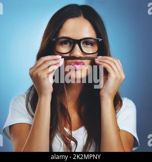 Montrant son côté original. Portrait en studio d'une jeune femme attirante en train de jouer une moustache avec ses cheveux sur fond bleu. Banque D'Images