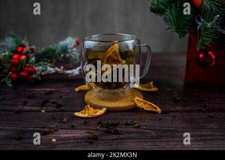 Une tasse de thé transparente est placée sur une table sombre, sur un support en bois, des tranches d'orange séchées flottent dans la tasse, des tranches d'agrumes se trouvent à côté, du thé est dispersé, une photo dans une touche sombre Banque D'Images