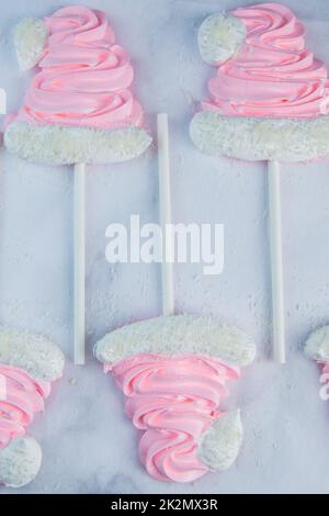 Meringue sous forme de chapeaux de Noël roses avec pompon et un revers blanc sur un bâton se trouvent sur parchemin blanc en deux rangées Banque D'Images