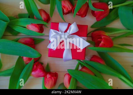 Des tulipes roses avec des feuilles vertes se trouvent dans un cercle sur fond de bois, au milieu du cadeau de Lesha dans un emballage rouge avec une boucle blanche Banque D'Images