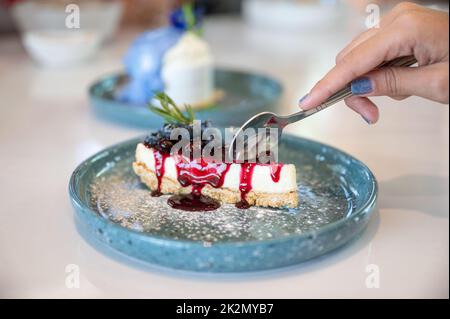 Main femelle à l'aide d'une cuillère coupant la tarte au fromage myrtille sur une plaque en céramique Banque D'Images