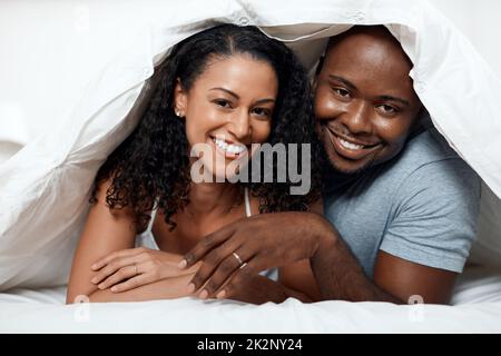 Toujours de bonne humeur ensemble. Portrait d'un jeune couple gai allongé dans un lit sous une couverture tout en regardant l'appareil photo à la maison le matin. Banque D'Images