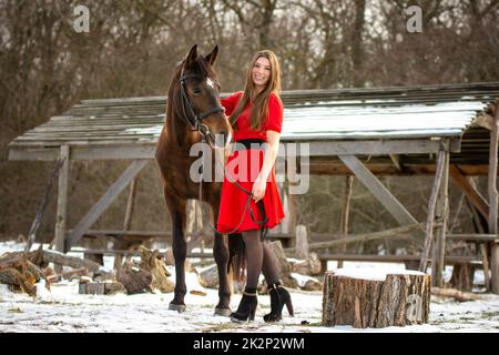 Une belle fille en robe rouge marche avec un cheval dans les rayons du soleil couchant sur fond de ruines anciennes Banque D'Images