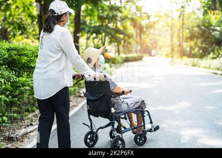 Aide-soignant et soins asiatique senior ou âgée vieille femme patiente assise en fauteuil roulant et portant un masque facial pour voyager dans le parc Banque D'Images