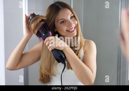 Portrait d'une jeune femme utilisant un sèche-cheveux à brosse ronde pour coiffer les cheveux au miroir de manière facile à la maison Banque D'Images