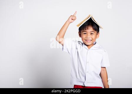 Asiatique adorable tout-petit souriant heureux portant étudiant thaïlandais uniforme pantalon rouge stand tenant le livre sur la tête comme le toit Banque D'Images