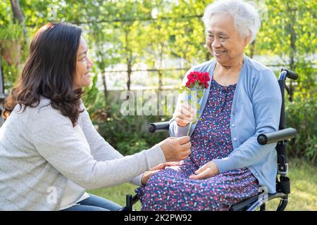 La fille du soignant se câlin et aide la vieille femme asiatique âgée ou âgée qui tient une rose rouge sur un fauteuil roulant dans le parc. Banque D'Images