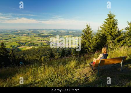 Une femme assise sur un banc, regardant du mont Hohenbogen à Neukirchen Heiligblut, une petite ville de la forêt bavaroise. Banque D'Images