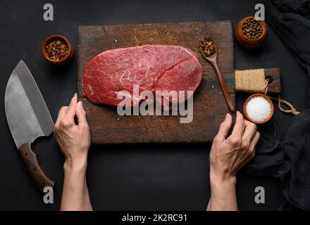 Le filet de bœuf cru se trouve sur une planche à découper en bois et des épices pour cuisiner sur une table noire, une vue sur le dessus et un grand couteau de cuisine Banque D'Images