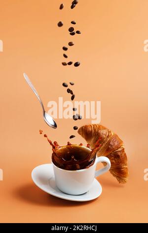 Tasse de café blanc tombant sur fond de table beige, éclaboussures, haricots, croissant Banque D'Images