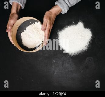 deux mains femelles tiennent un tamis rond en bois avec de la farine de blé sur une table noire, vue de dessus Banque D'Images