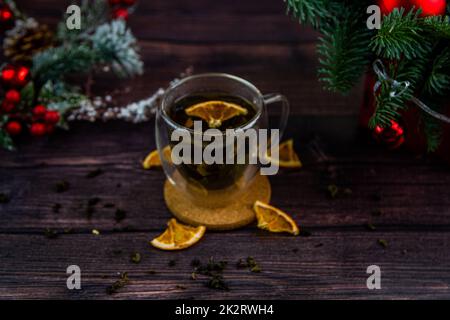 Une tasse de thé transparente est placée sur une table sombre, sur un support en bois, des tranches d'orange séchées flottent dans la tasse, des tranches d'agrumes se trouvent à côté, du thé est dispersé, une photo dans une touche sombre Banque D'Images