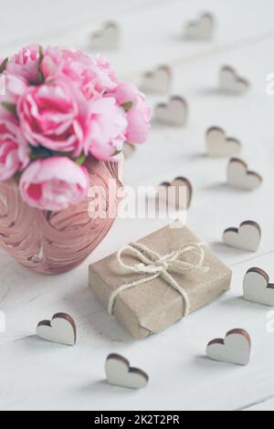 Belle composition avec cadeau enveloppé dans du papier kraft et de délicates roses dans un vase rose sur fond de bois blanc Banque D'Images