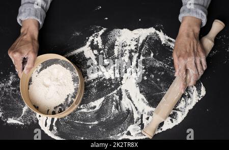 Les mains des femmes tiennent un tamis rond en bois avec de la farine de blé blanc sur une table noire, vue de dessus Banque D'Images