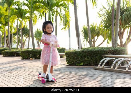 Joyeux asiatique petit enfant fille porter un casque sûr jouant de la planche à pied rose sur la route dans le parc à l'extérieur le jour d'été Banque D'Images