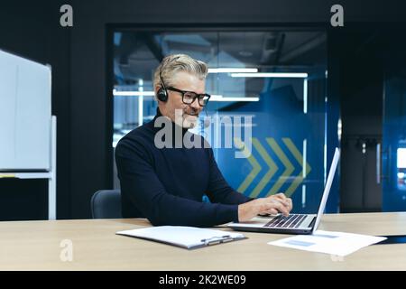 Un investisseur senior aux cheveux gris travaillant dans un bureau moderne à l'aide d'un ordinateur portable et d'un micro-casque pour les appels vidéo et les réunions à distance avec ses collègues et partenaires. Banque D'Images