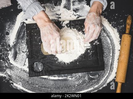 deux mains de femmes pétrient la pâte de farine de blé blanc sur une table noire, vue de dessus Banque D'Images