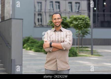 Jeune homme d'affaires réussi souriant et regardant la caméra avec des bras croisés, homme à l'extérieur du bâtiment de bureau portant une chemise et des lunettes, startup investisseur. Banque D'Images