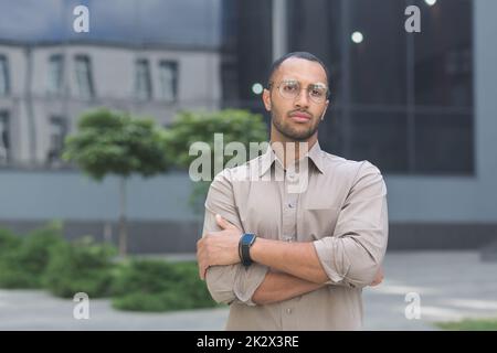 Sérieux et pensif jeune employé de bureau avec les bras croisés regardant la caméra, homme d'affaires dans la chemise à l'extérieur du bâtiment de bureau pensif. Banque D'Images