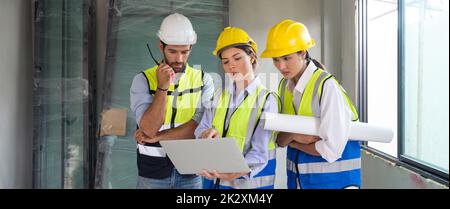 Un jeune architecte pointant vers un moniteur d'ordinateur portable explique au contremaître et à l'ingénieur un plan d'étage. Tout le monde porte un casque et un gilet de sécurité. Milieu de travail au chantier du projet de logement. Banque D'Images