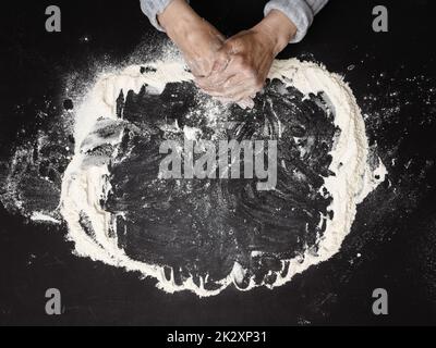 Farine de blé blanc tamisée sur une table noire et deux mains femelles, vue du dessus. Cuisiner à la maison Banque D'Images